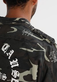 Giacca con stampa mimetica in verde scuro e nero, con testo grafico bianco e dettagli logori. Colletto chiuso e dettaglio con bottone sulla spalla.