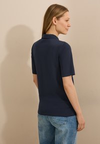 Donkerblauwe polo met korte mouwen en een kraag, gemaakt van zacht materiaal. Het shirt heeft een getailleerde pasvorm en wordt gedragen met lichtblauwe jeans.