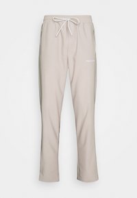 Beige tepláky s elastickým pasem, šňůrkou na stažení a bočními zipovými kapsami. Na levém boku mají potisk loga. Hladká struktura látky.
