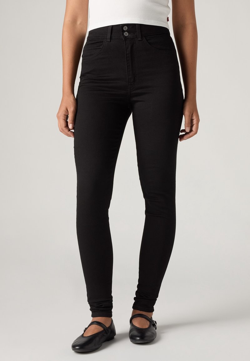 Femmes portant un jean skinny noir taille haute associé à des chaussures Mary Jane noires et un top blanc sans manches, debout devant un fond uni.