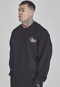 Felpa di un blu navy scuro con scollatura a girocollo, vestibilità oversize e logo bianco sul petto sinistro. Texture liscia, polsini e orlo a costine.
