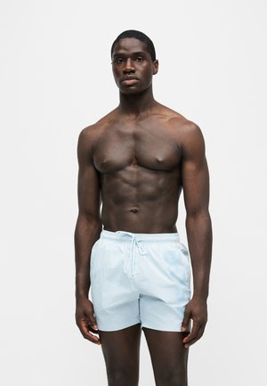 Muskelbepackter Mann, der frontal steht und hellblaue Badeshorts mit Kordelzug vor einem schlichten weißen Hintergrund trägt.