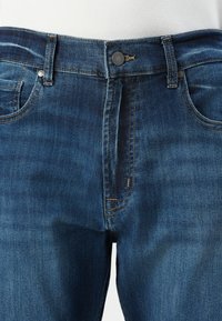 Jeans de denim azul con diseño clásico de cinco bolsillos, con botón visible y pasadores para cinturón, combinados con una camiseta blanca.