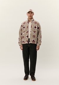 Mladi moški, oblečen v bež vzorčasti flis jakni, črne kavbojke, temno rjave čevlje in kremno barvno kapo z rdečimi črkami.