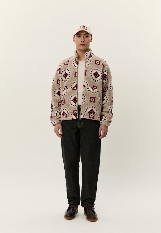 Mladi moški, oblečen v bež vzorčasti flis jakni, črne kavbojke, temno rjave čevlje in kremno barvno kapo z rdečimi črkami.