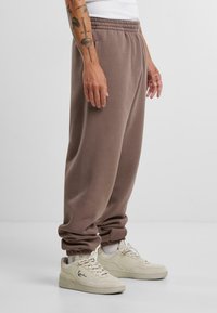 Brązowe bawełniane dresy z elastycznym pasem i zwężanymi mankietami, połączone z kremowymi sneakersami. Posiada widoczne szwy i luźny krój.