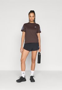 Brązowy sportowy t-shirt z niebieskimi paskami na rękawach, zestawiony z czarnymi szortami. Model trzyma czarną butelkę na wodę, ma na sobie czarne trampki i białe skarpetki.