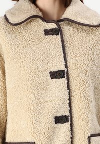 Beige shearling-takki, jossa on teksturoitu pinta. Siinä on pyöreä kaulus, tummat reunukset ja kolme mustaa nappia edessä.