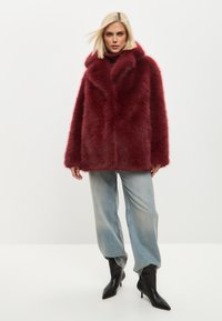 Abrigo de piel sintética burdeos con cuello alto, que presenta una silueta rectangular y mangas amplias, combinado con vaqueros azul claro y botas negras de tacón.
