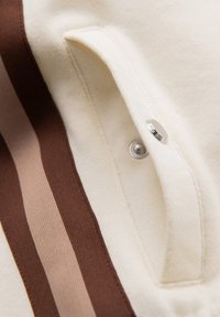 Gros plan d’un tissu crème avec un bouton-pression métallique et trois bandes verticales en brun foncé, brun moyen et beige clair.