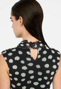 Schwarzes ärmelloses Kleid mit großen weißen Punkten, das über einen Schlüssellochausschnitt und zwei schwarze Knöpfe auf der Rückseite verfügt, aus strukturiertem Stoff.