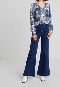 Blouse à manches longues et col en V en patchwork bleu et blanc avec des motifs floraux, associée à un jean en denim foncé évasé et des bottines blanches à talons.