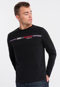 Schwarzes Langarm-T-Shirt mit dem Text "OMBRE MENSWEAR" in fettem Weiß und Rot, versehen mit horizontalen farbigen Streifen auf der Brust.
