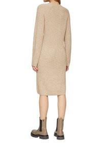Beige gebreide jurk met lange mouwen en een aansluitende pasvorm, met een ronde halslijn en een lengte tot halverwege de scheen. Gedragen met bruine enkellaarsjes.