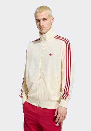 Jeune homme aux cheveux blonds courts portant une veste de survêtement crème avec des rayures rouges et un pantalon rouge Adidas, debout les mains dans les poches.