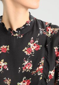 Robe noire à fleurs avec des roses rouges et jaunes, encolure à volants, et motif floral délicat, visible jusqu'à la zone des épaules.