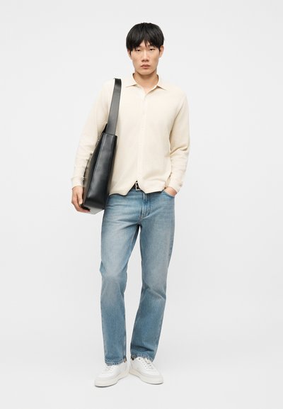 Jeune homme portant une chemise crème à boutons, un jean bleu clair, des baskets blanches, et portant un grand sac fourre-tout en cuir noir sur l'épaule.