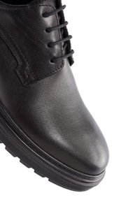 Chaussure noire en cuir à lacets, avec bout arrondi et semelle épaisse rainurée, montrée en gros plan sous un angle supérieur latéral.