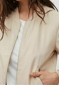 Béžová lehká bomber bunda se zipovým uzávěrem, žebrovaným l�ímcem a bočními kapsami. Hladká textura, minimalistický design a žádné viditelné vzory.