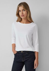 Jonge vrouw met lang blond haar, gekleed in een witte shirt met driekwart mouwen en donkerblauwe jeans, staande tegen een effen grijze achtergrond.