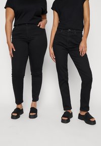 Dois pares de jeans de denim preto em modelos, um justo com comprimento até ao tornozelo e o outro de corte reto. Ambos usados com camisetas pretas de mangas curtas.