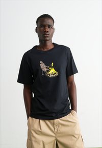 Camiseta de algodón negra con un gráfico de un esqueleto sentado junto a una fogata, con llamas amarillas vibrantes. Manga corta y corte relajado.