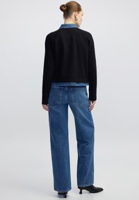 Zwarte geribbelde jas met een denim kraag, gecombineerd met high-waisted blauwe jeans. De jeans hebben een losse pasvorm en rafelige zomen, aangevuld met zwarte hakken.