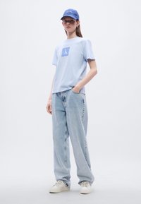 Ανοιχτό μπλε βαμβακερό μπλουζάκι με τετράγωνο λογότυπο, συνδυασμένο με ξεθωριασμένα ανοιχτό μπλε τζιν και λευκά αθλητικά παπούτσια. Φορά επίσης μπλε καπέλο.