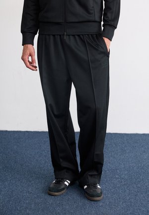 Person trägt schwarze Adidas-Trainingshose, schwarze Zip-Up-Jacke und schwarze Sneakers mit weißen Streifen, steht auf blauem Teppich.