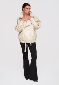 Femme ajustant le col d'une veste en cuir crème surdimensionnée, portant un pantalon noir évasé et des chaussures noires pointues, debout sur un fond uni.