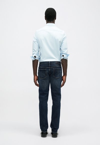 Mann stående med ryggen mot, iført en lyseblå langermet skjorte med oppbrettede ermer, mørkeblå jeans og svarte sko, mot en ensfarget bakgrunn.