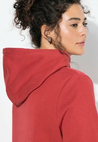 Czerwona bluza z kapturem o miękkiej fakturze, charakteryzująca się luźnym krojem i minimalistycznym wzornictwem. Akcesorium to pojedyncza kolczyk-koło.