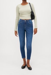 Blusa de manga larga verde claro combinada con jeans ajustados azules y bailarinas negras. Los jeans tienen un diseño clásico de cinco bolsillos y cintura alta.