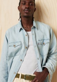 Light blue denim shirt with buttoned chest pockets, worn over a white ribbed tank, paired with beige pants and a green belt. Smooth texture.