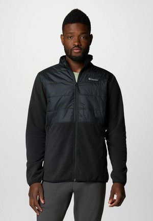 Veste noire pour homme avec un buste matelassé, fabriquée en matériau en polaire doux. Elle présente une fermeture éclair à l'avant, un col haut et un logo discret sur la poitrine.