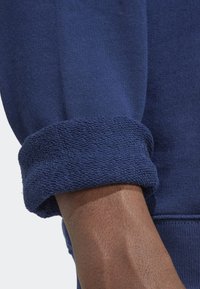 Puño de sudadera azul marino hecho de algodón texturizado, con un dobladillo enrollado y tela suave. Acercamiento al brazo mostrando el tono de piel.