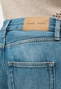 Κοντινή λήψη πίσω μέρους μπλε τζιν τζιν με ταμπέλα από ταμπά δέρμα και μερικό μέρος από μπεζ πλεκτό τοπ ορατό.