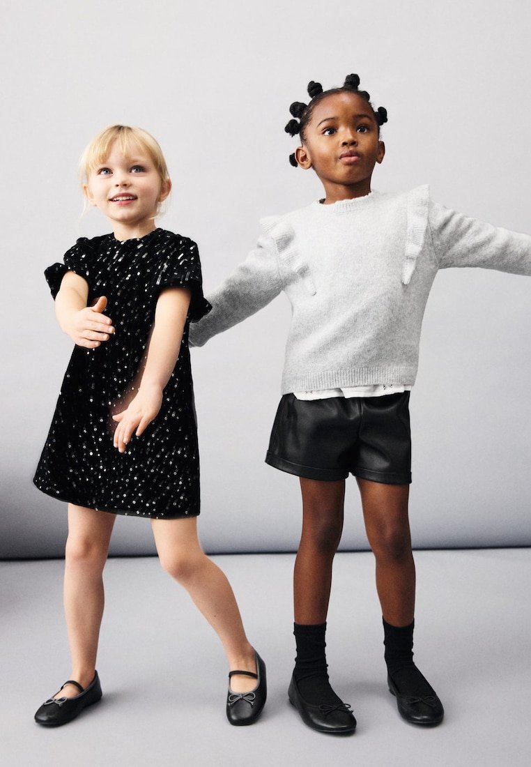 Dvě dívky stojí s nataženýma rukama; jedna má na sobě černé šaty s flitry, druhá světlý šedý svetr a černé šortky.