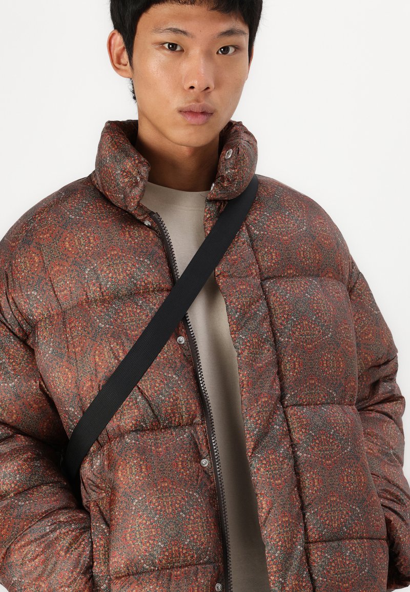 Gepolsterte Steppjacke in Rostfarbe mit geometrischem Muster, hohem Kragen, Reißverschluss und Druckknöpfen. Über einem beigen Oberteil getragen.