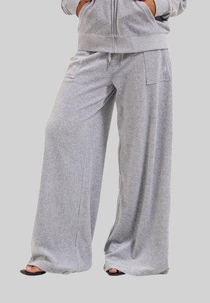 Vrouw draagt loszittende lichtgrijze joggingbroek met brede pijpen en een bijpassende zip-up hoodie, staand met handen in de zakken.