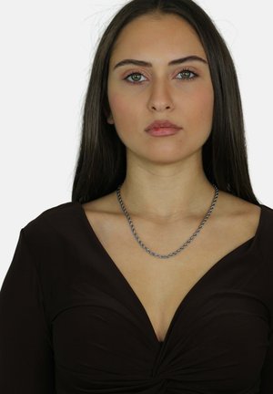 Collana in argento con maglie attorcigliate, indossata da un modello con una maglietta marrone a profondissimo scollo a V, caratterizzata da un tessuto liscio e un design aderente.