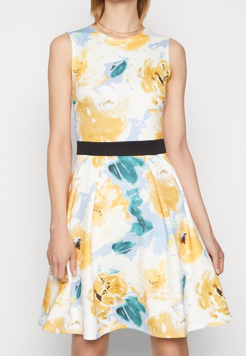 Robe sans manches avec motif floral jaune et blanc, feuilles vertes, fond bleu clair, taille ajustée avec ceinture noire, jupe longueur genou.