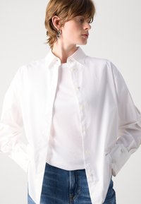 Hvit langermet skjorte med krage som kan kneppes, med en sprø tekstur og litt oversized passform, båret over en enkel hvit topp og blå jeans.