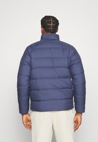 Veste matelassée bleu marine avec des coutures horizontales, col montant et une coupe décontractée. Fabriquée en matériau lisse et isolant, adaptée aux temps froids.