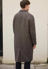 Mees seljaga, kes kannab pikka pruuni tekstuuriga üleriiet ja tumedaid pükse, seisab õues beežist seinast ja sillutisest vastu.