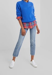 Person klädd i blå tröja över röd rutiga skjorta, ljusblå cropped jeans och vita sneakers står på grått golv.