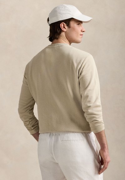 Camisa bege de manga comprida com decote redondo e punhos canelados, combinada com calças brancas. A camisa tem uma textura suave e design minimalista.