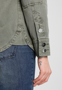 Oliivivihreä takki jämäkkällä muotoilulla, jossa on hopeiset nappikiinnitykset hihassa, yhdistettynä sinisiin farkkuihin ja näkyvään tatuointiin ranteessa.