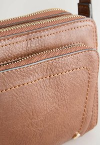 Sac en cuir marron avec trois compartiments zippés. Surface texturée et détails en fil doré contrastant sur les zips et les coutures. Design compact.