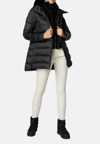 Schwarzer Steppmantel mit Kapuze, gesteppte Optik und Rei ßverschlusstaschen. Getragen über einem schwarzen Pelztop und weißen Leggings, kombiniert mit schwarzen Stiefeln.